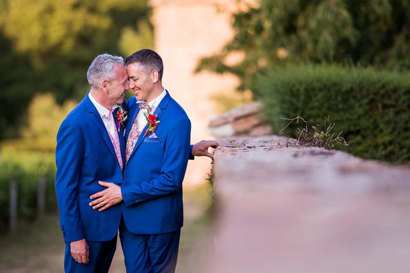 photo de mariage d'un couple d'hommes posés sur un muret portant chacun un costume bleu